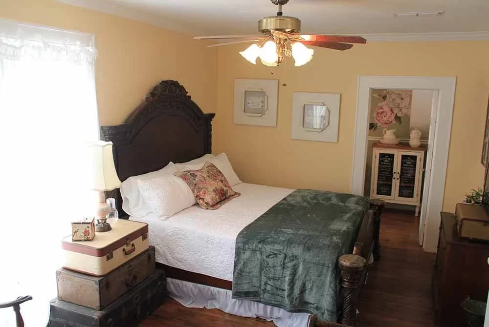 A bright bedroom with soft yellow walls, a dark wood bed with a green velvet throw, a ceiling fan, and an open doorway to a vanity area.