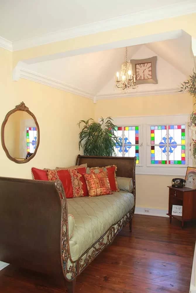 A bright seating area with a wooden sleigh daybed against yellow walls, featuring an oval mirror, an antique phone, and colorful stained glass windows.