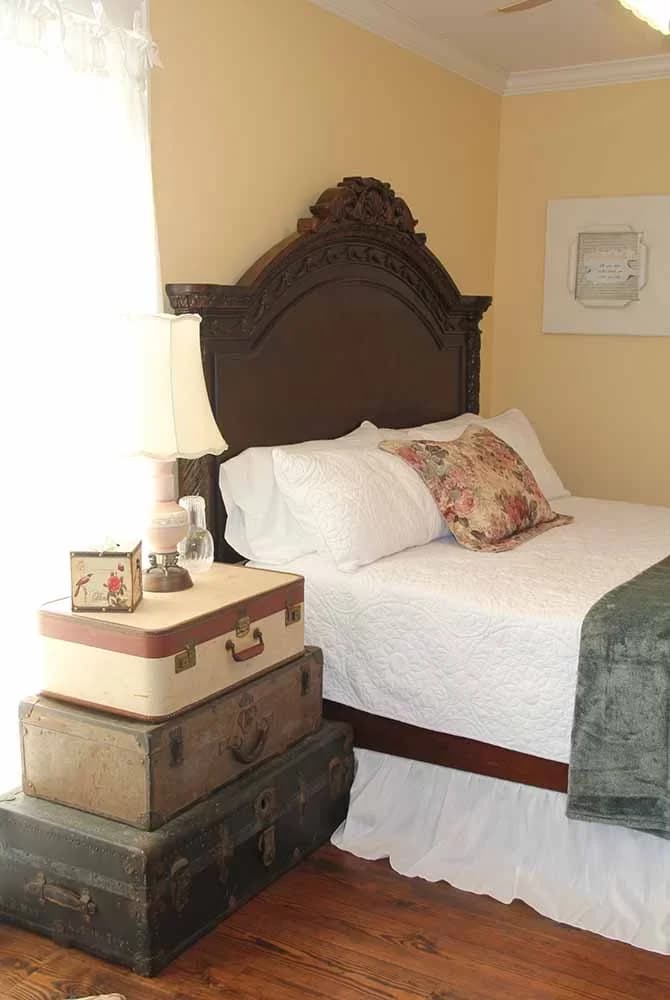 A bright bedroom with soft yellow walls, a dark wood bed, and a unique nightstand made of stacked vintage suitcases topped with a lamp.