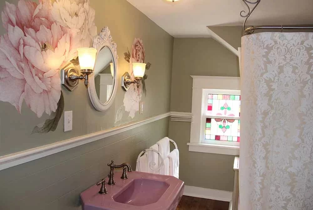 A whimsical bathroom with pink floral details, a pink-topped sink with vintage fixtures, a bidet, and a window with stained glass.