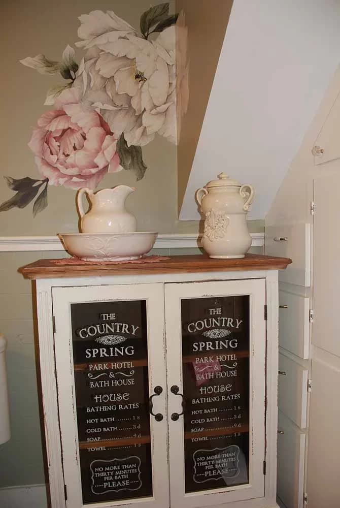 A charming white cabinet with chalkboard-style doors, topped with a pitcher and basin, set against a wall with a large pink floral detail.