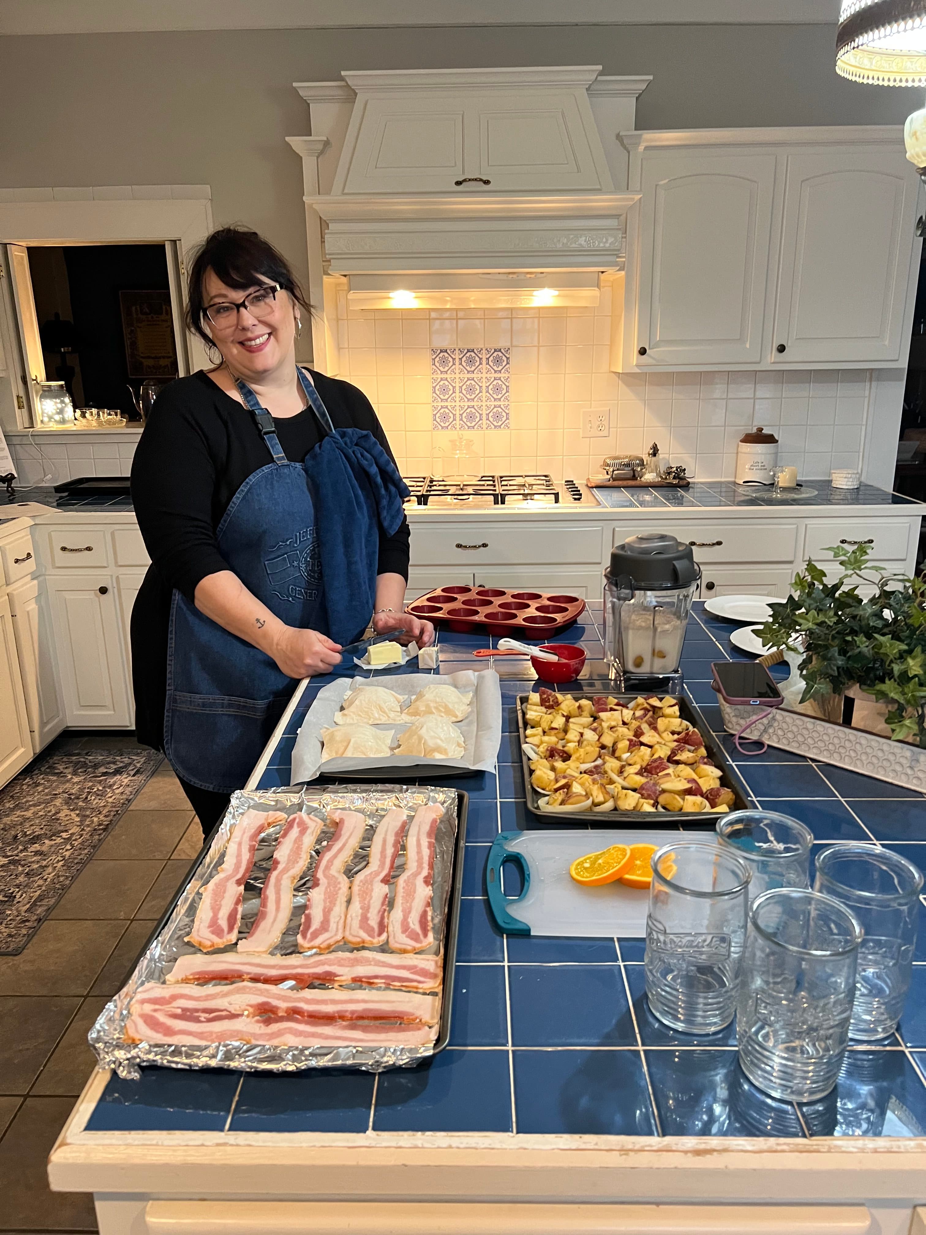 A smiling woman in an apron prepares food in a bright kitchen, surrounded by ingredients and cookware.