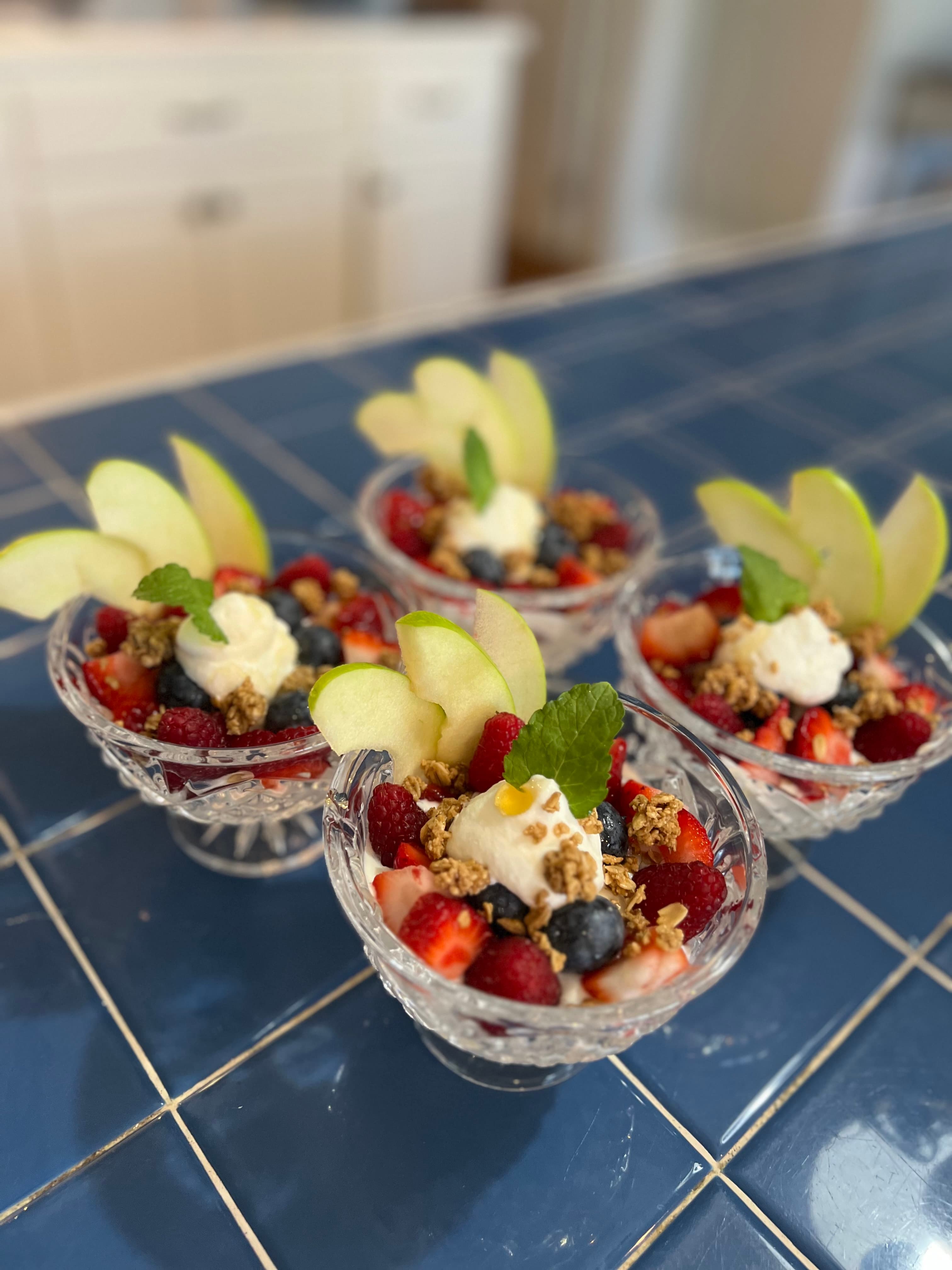 Four glass bowls of mixed berry dessert topped with granola, whipped cream, and apple slices on a blue tiled surface.