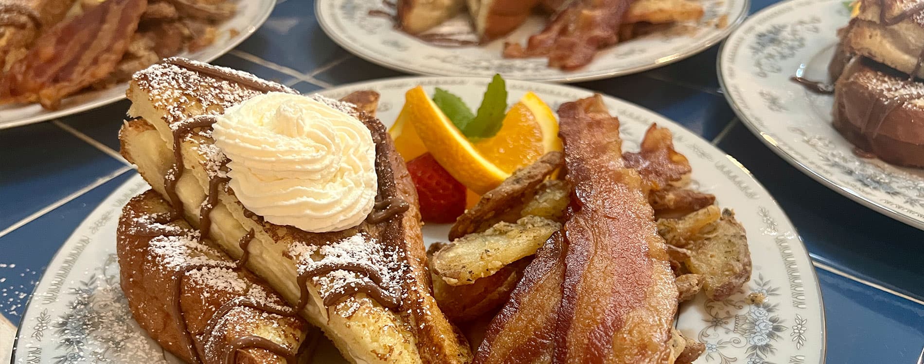 A plate of French toast with whipped cream, beside crispy bacon and garnished with fresh fruit.