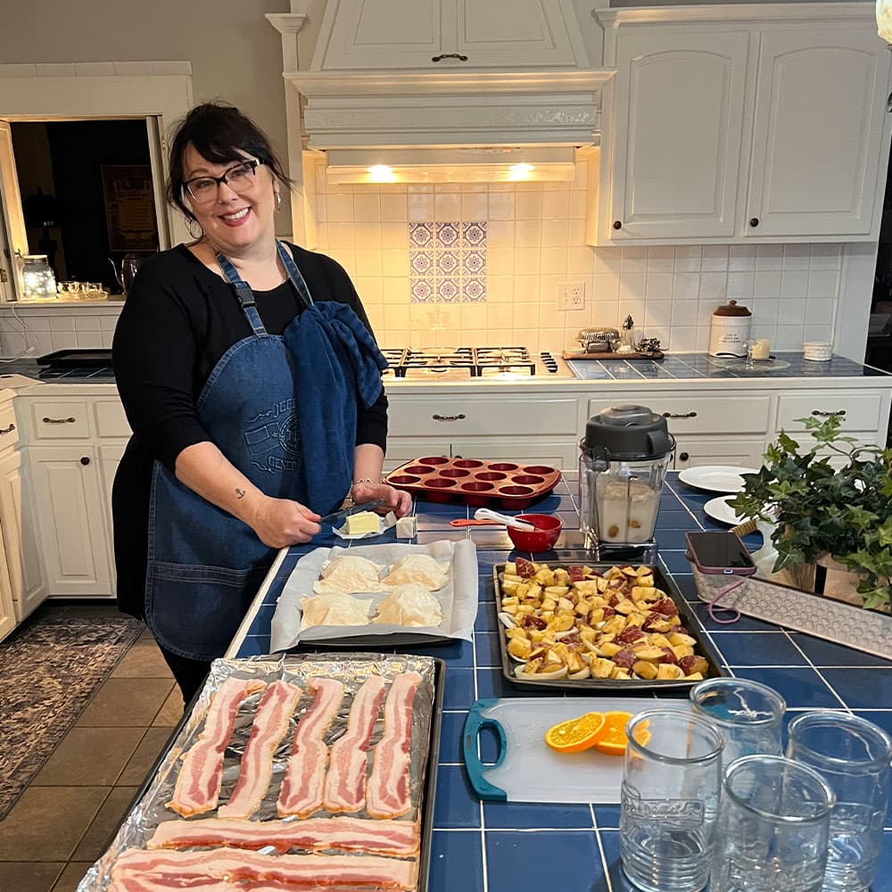 A smiling woman in an apron prepares food in a bright kitchen filled with various cooking ingredients.
