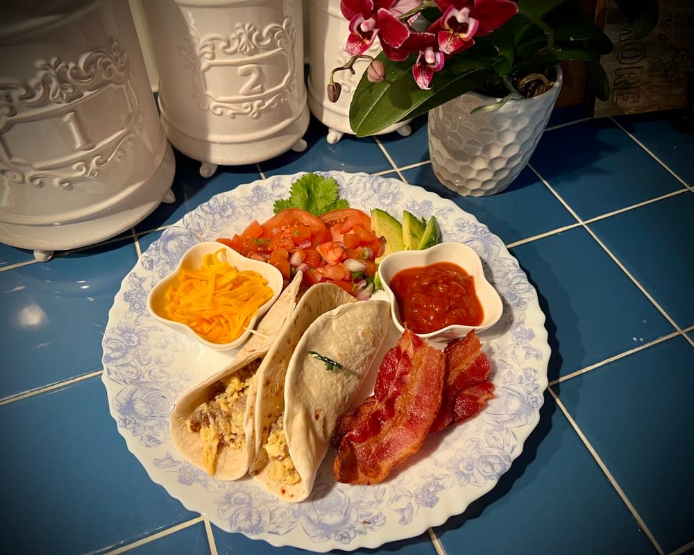 A plate of breakfast tacos with eggs, bacon, cheese, fresh tomatoes, lettuce, avocado, and salsa, garnished with an orchid plant in the background.