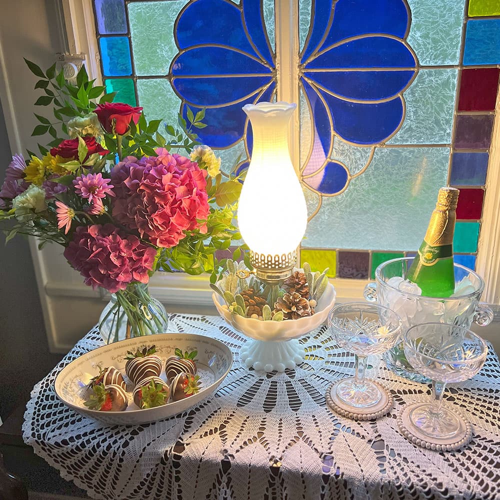 A table displays a bouquet of flowers, a glowing lamp, chocolate-covered strawberries, champagne, and crystal glassware against a colorful stained glass background.