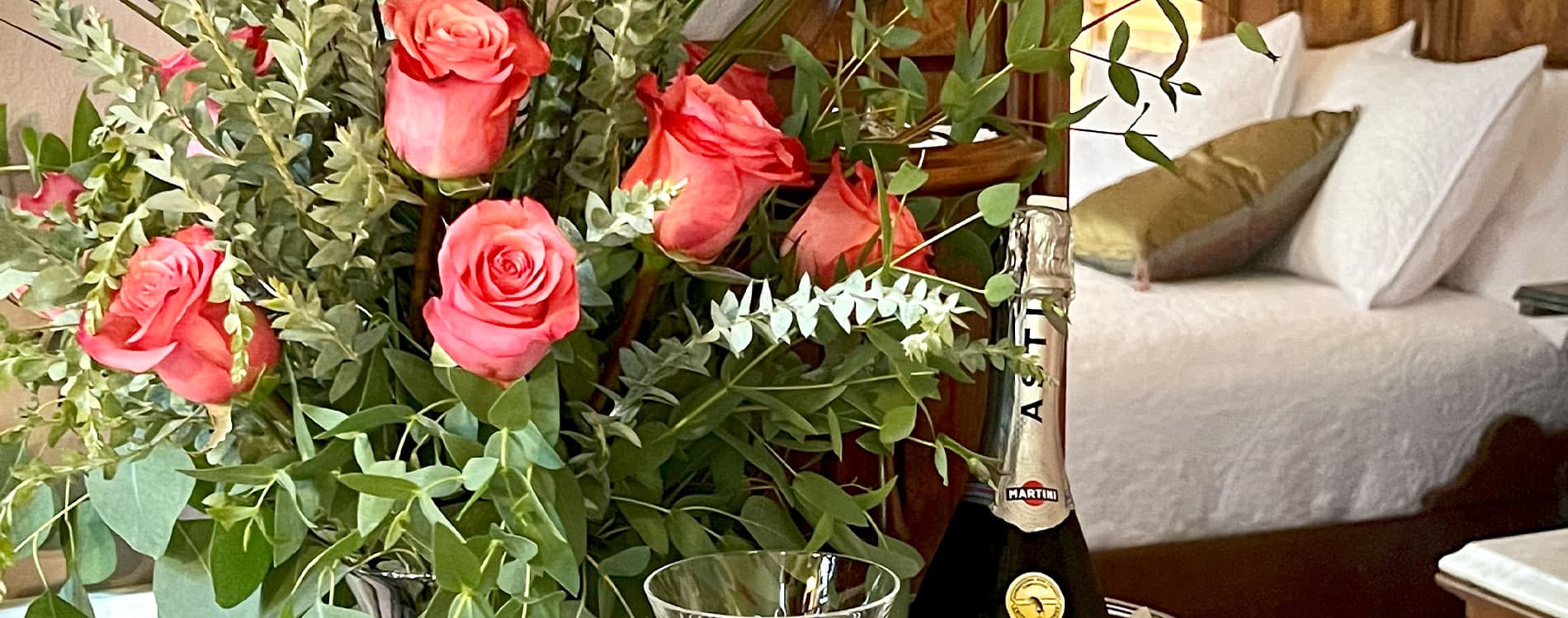 A bouquet of pink roses with greenery beside a bottle of sparkling wine and a glass, set against a cozy bedroom backdrop.