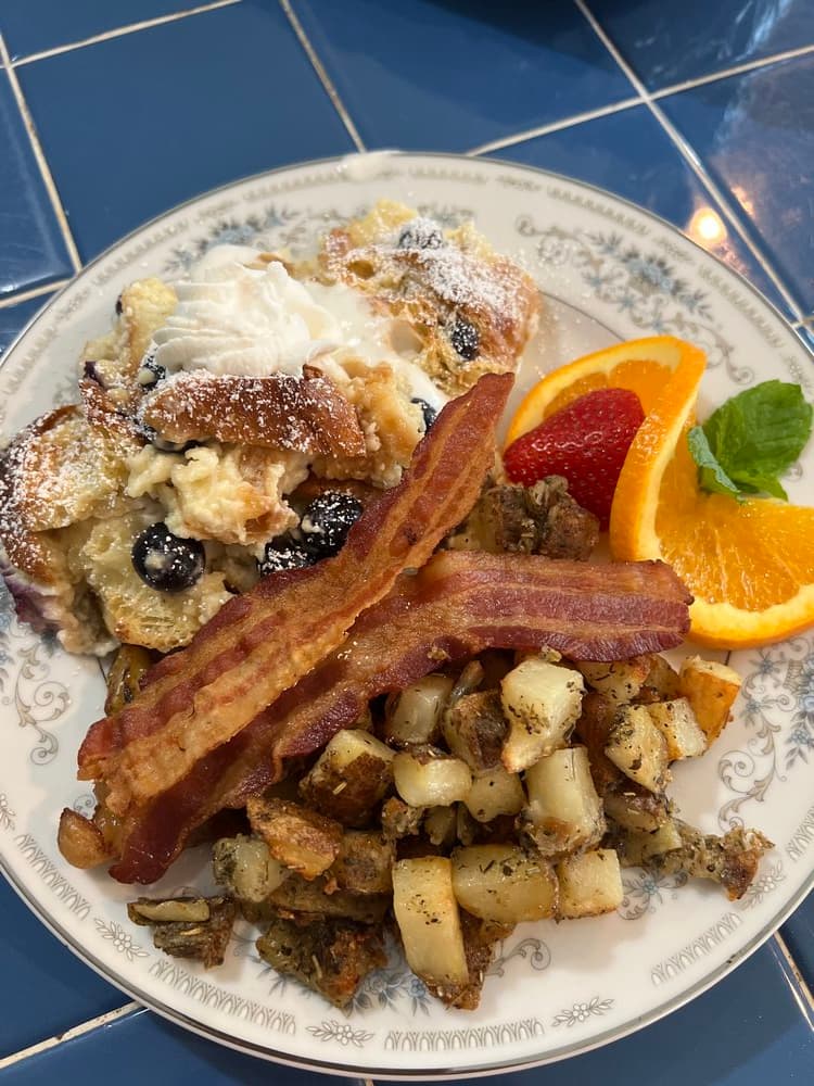 A plate of breakfast featuring a blueberry pastry, whipped cream, bacon, roasted potatoes, and orange and strawberry slices.
