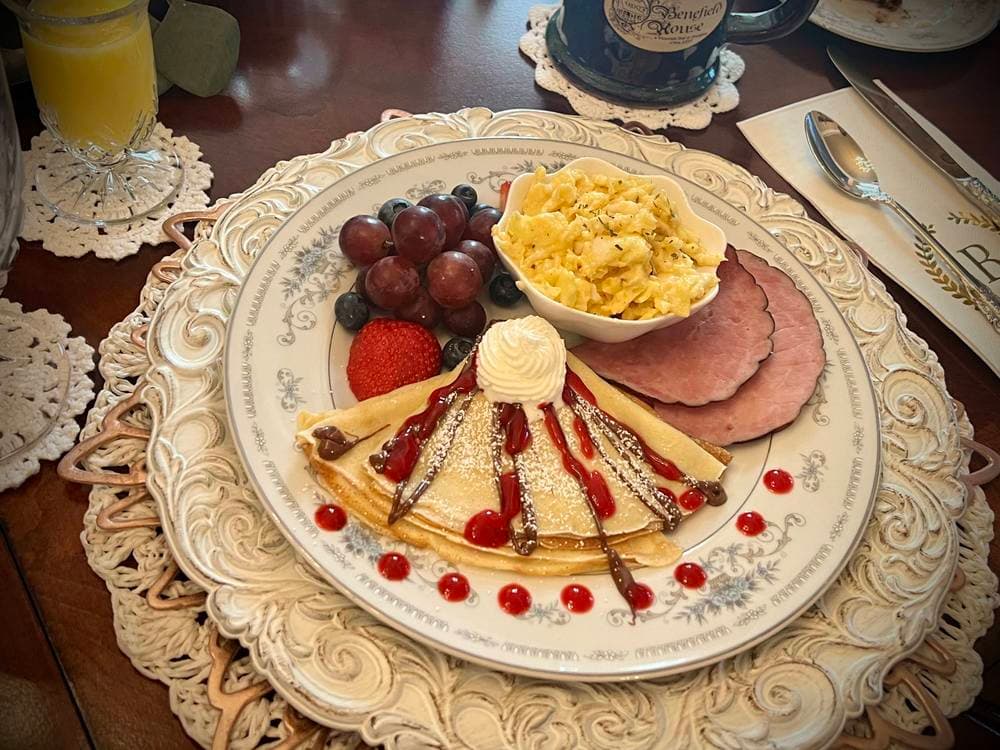 A beautifully arranged breakfast plate featuring a crepe topped with whipped cream and berries, scrambled eggs, ham, and assorted fruit.