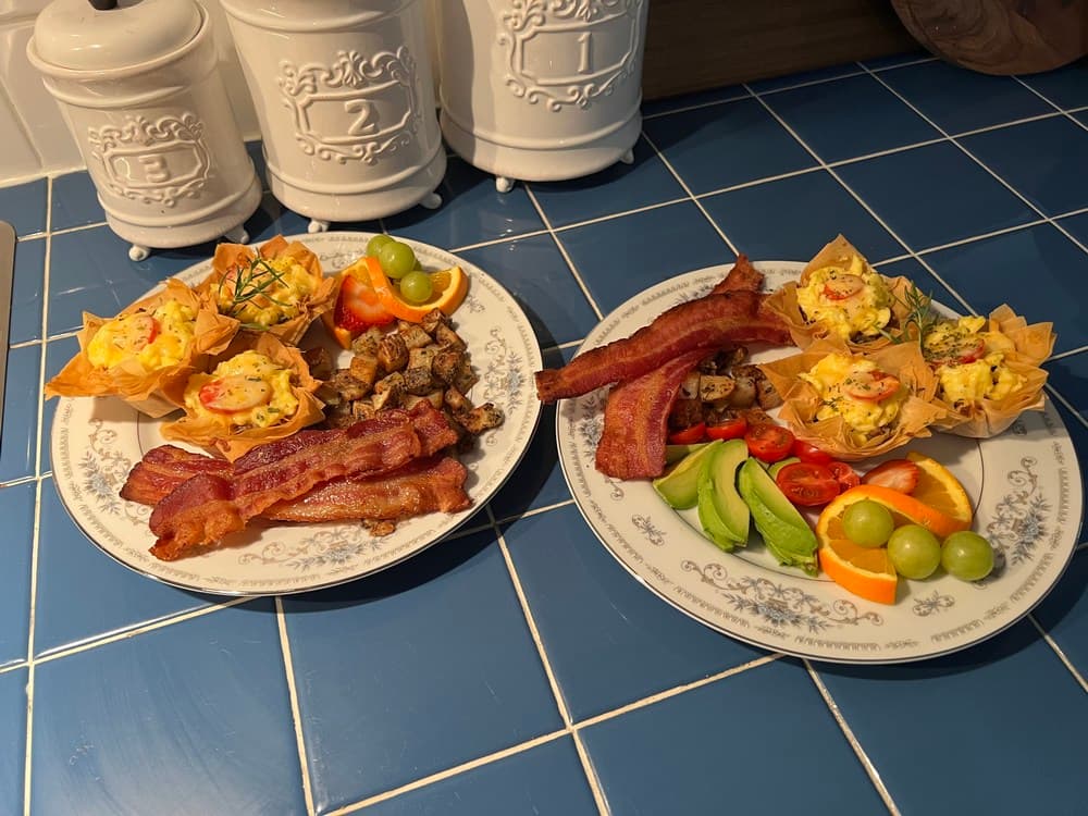 Two plates of breakfast featuring bacon, egg cups, vegetables, and fresh fruit on a blue tiled surface.
