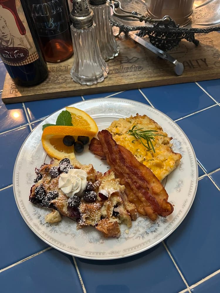 A plate of breakfast featuring blueberry pancakes with whipped cream, scrambled eggs, crispy bacon, and orange slices.