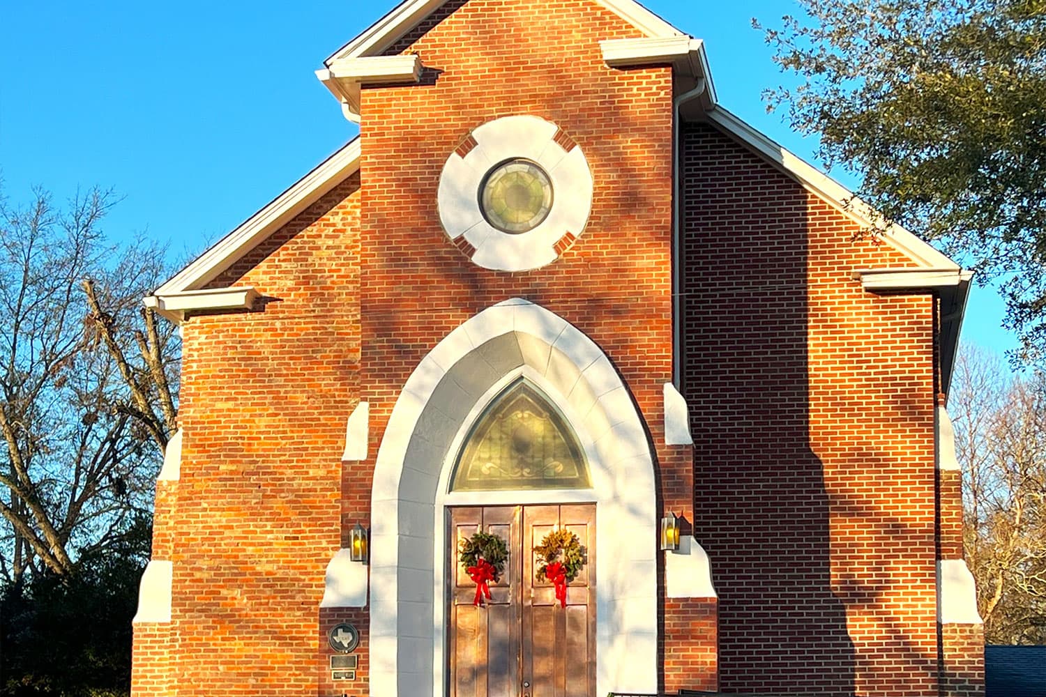 A brick church with a circular window and festive wreaths on the double doors.