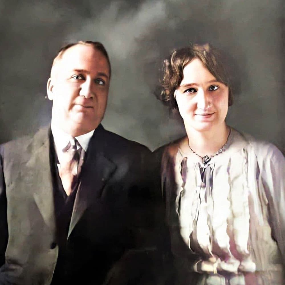 A man and woman are posed together, both dressed in early 20th-century attire, with a soft, blurred background.