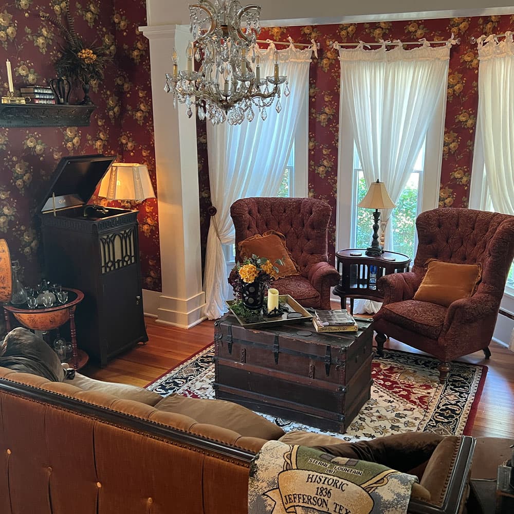 A cozy living room featuring vintage furniture, a decorative rug, and large windows with sheer curtains.