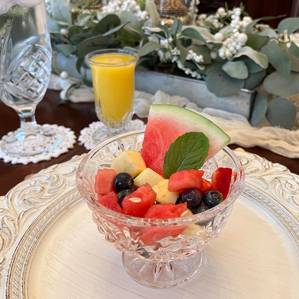 A decorative glass bowl filled with a colorful fruit salad featuring watermelon, blueberries, and other fruits, accompanied by a yellow drink and greenery in the background.