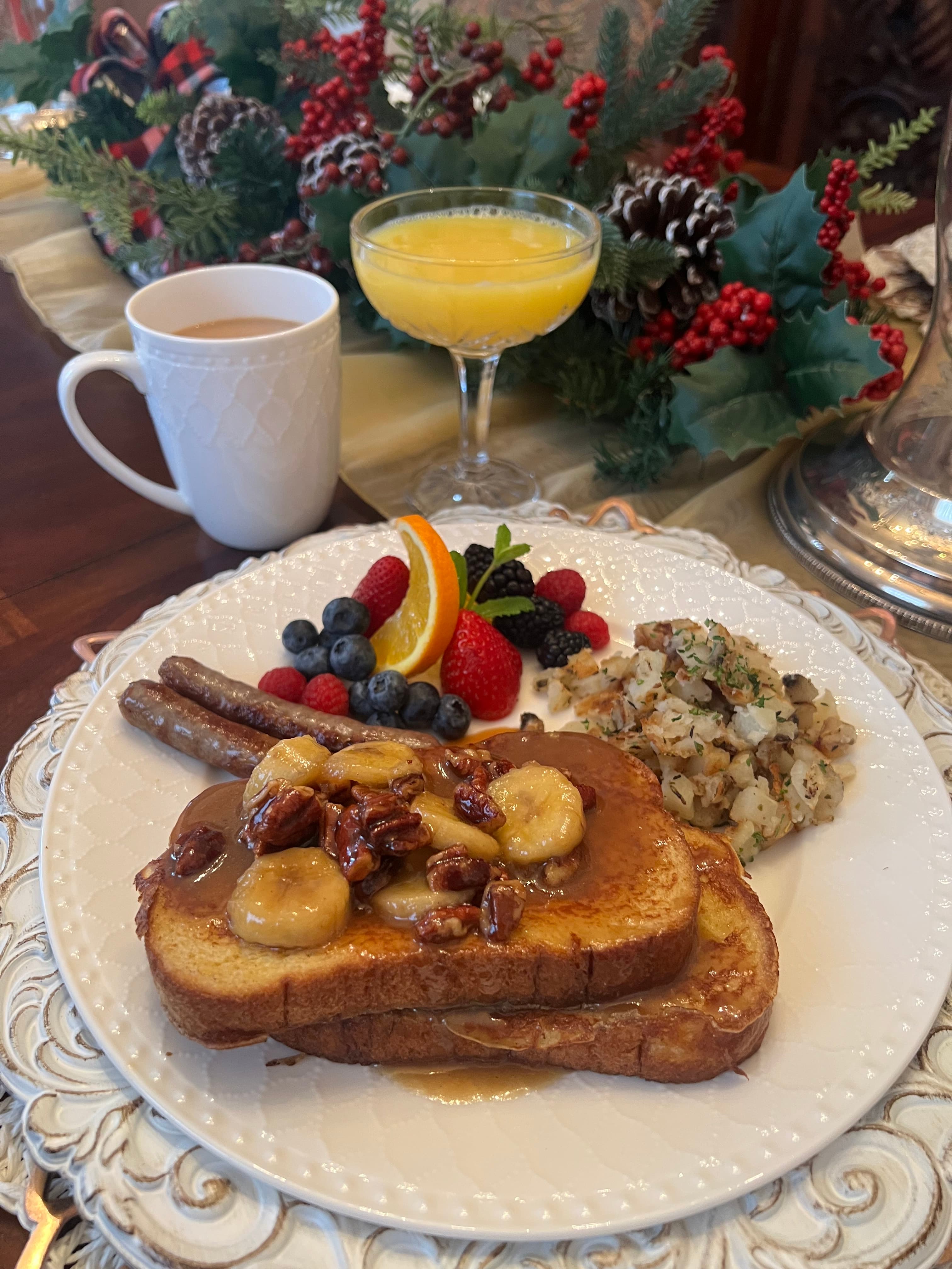 A festive breakfast platter featuring French toast topped with bananas and nuts, alongside sausage, mixed berries, orange slices, and a cup of coffee.
