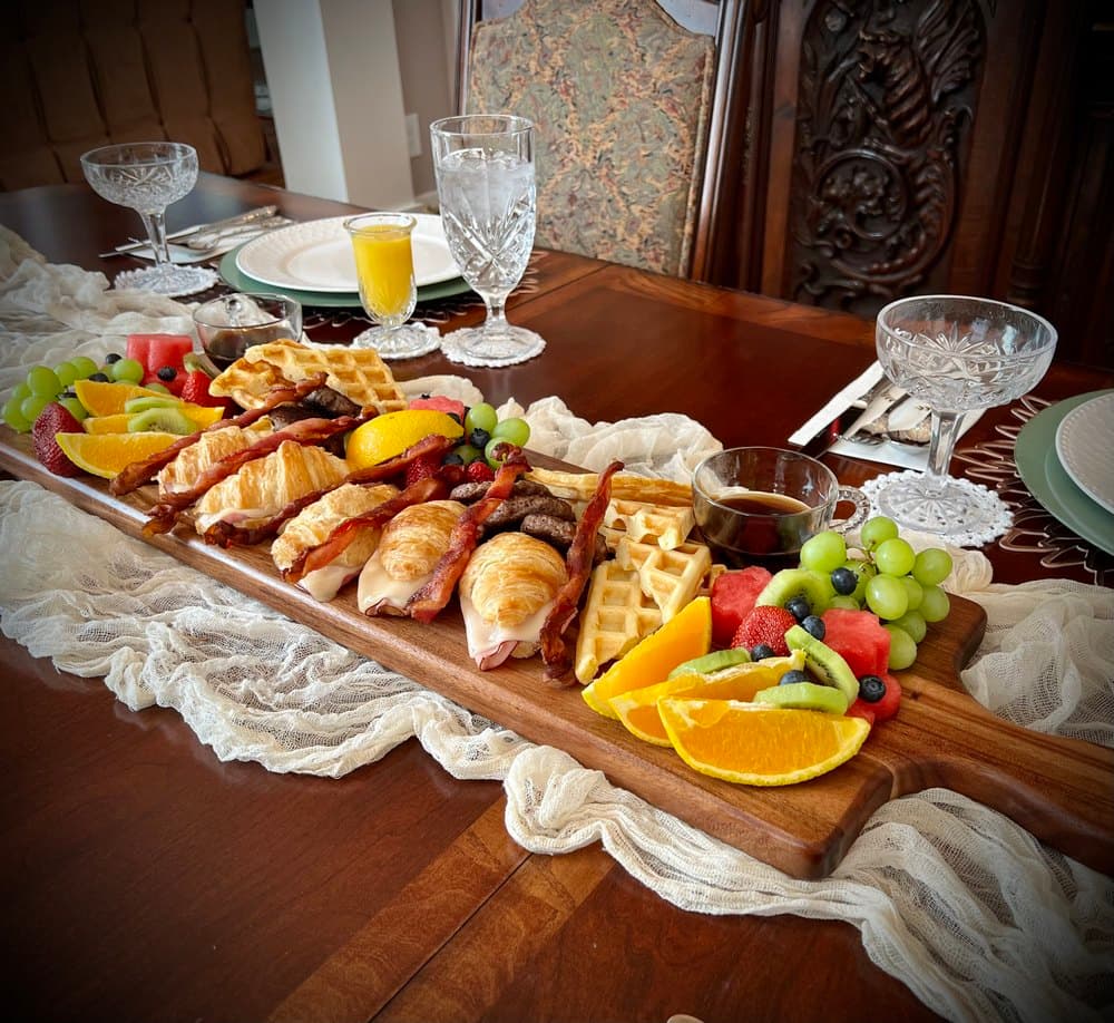 A beautifully arranged breakfast platter featuring waffles, chicken, bacon, assorted fruits, and syrup on a wooden board.