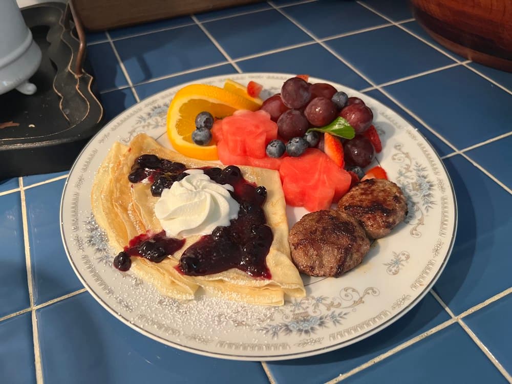 A plate of crepes topped with berry jam and whipped cream, accompanied by assorted fresh fruits and sausage patties.