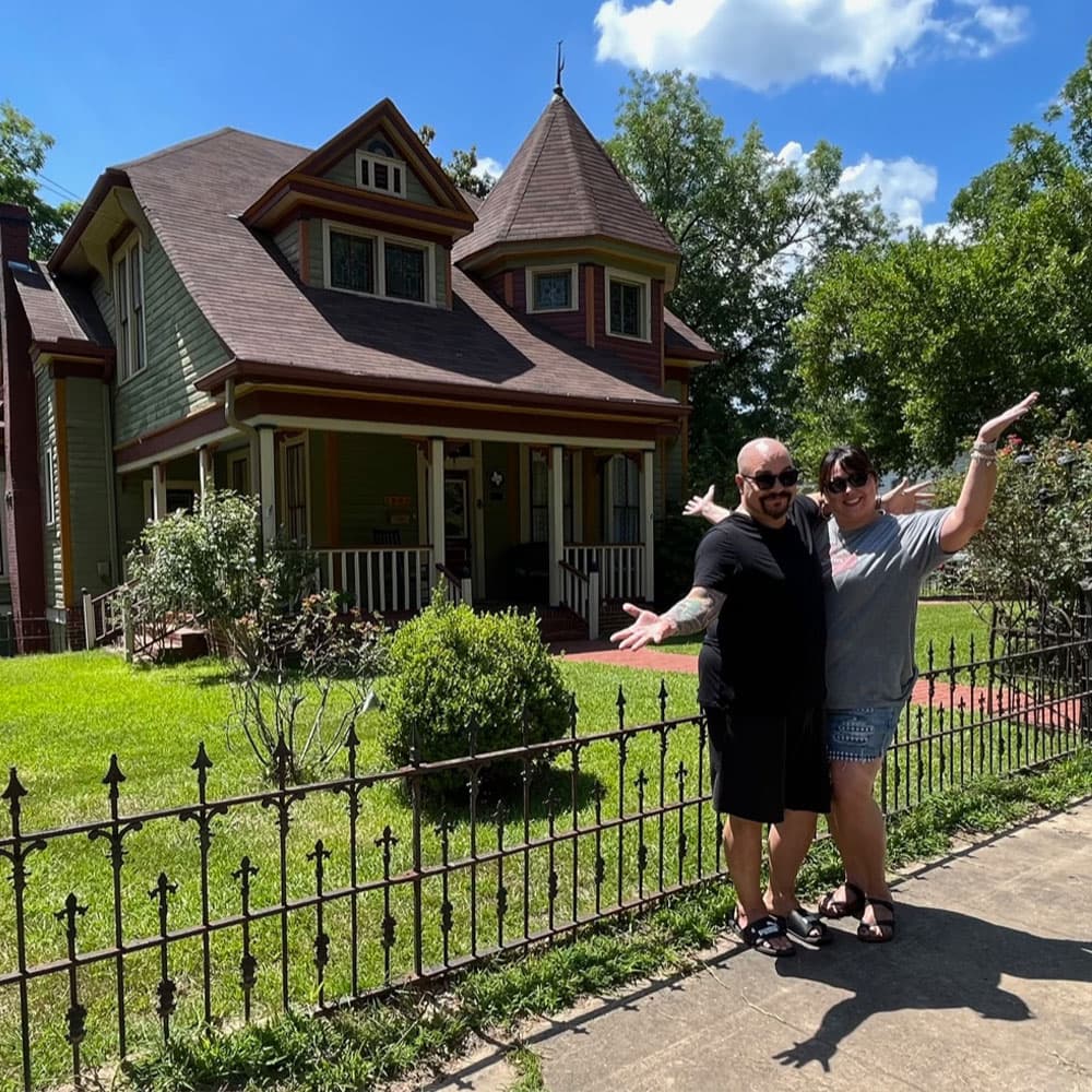A couple poses joyfully in front of a charming Victorian-style house.