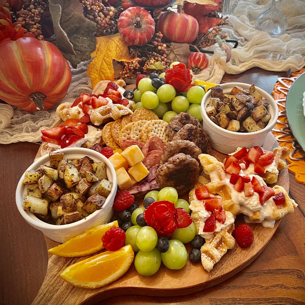 A colorful assortment of fruits, meats, cheeses, waffles, and roasted potatoes arranged on a wooden platter with autumn decorations in the background.