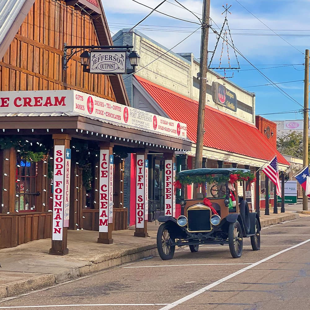 A vintage car is parked outside a quaint storefront adorned with festive decorations in a charming small town.