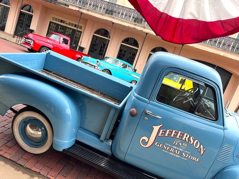 A vintage blue truck with "Jefferson Texas General Store" written on the side is parked in front of colorful classic cars and a building with ornate balconies.