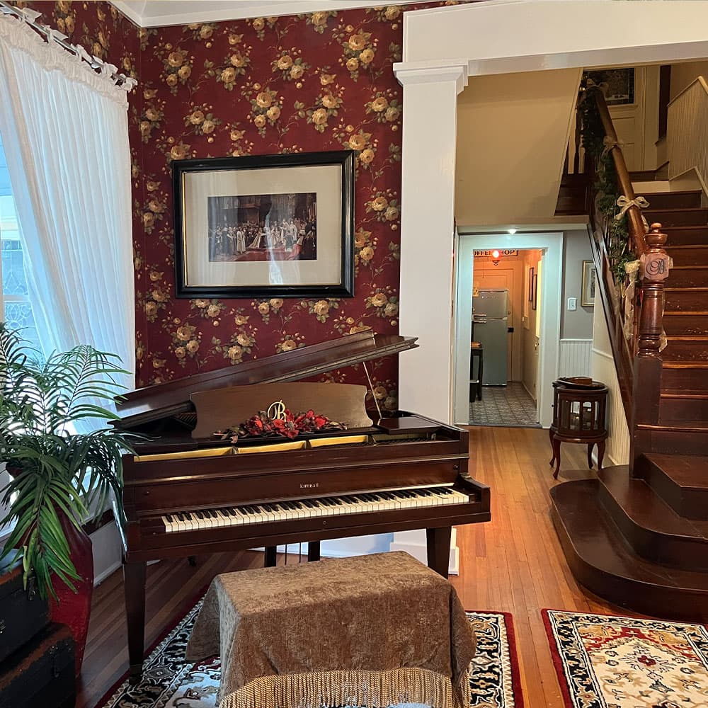 A grand piano sits in a warmly decorated room with floral wallpaper and wooden staircase in the background.