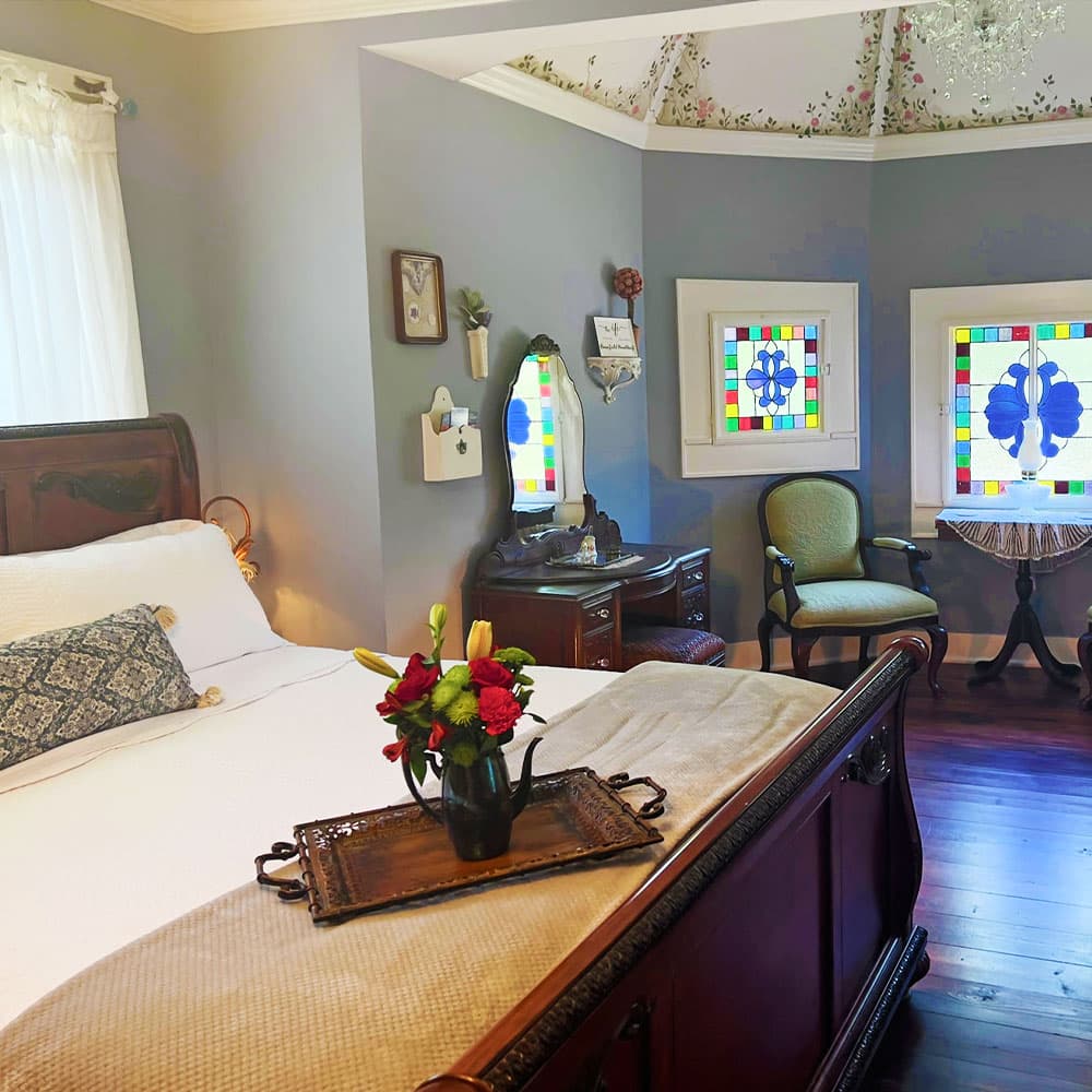 Cozy bedroom featuring a bed with decorative pillows, a vintage dresser, and stained glass windows.