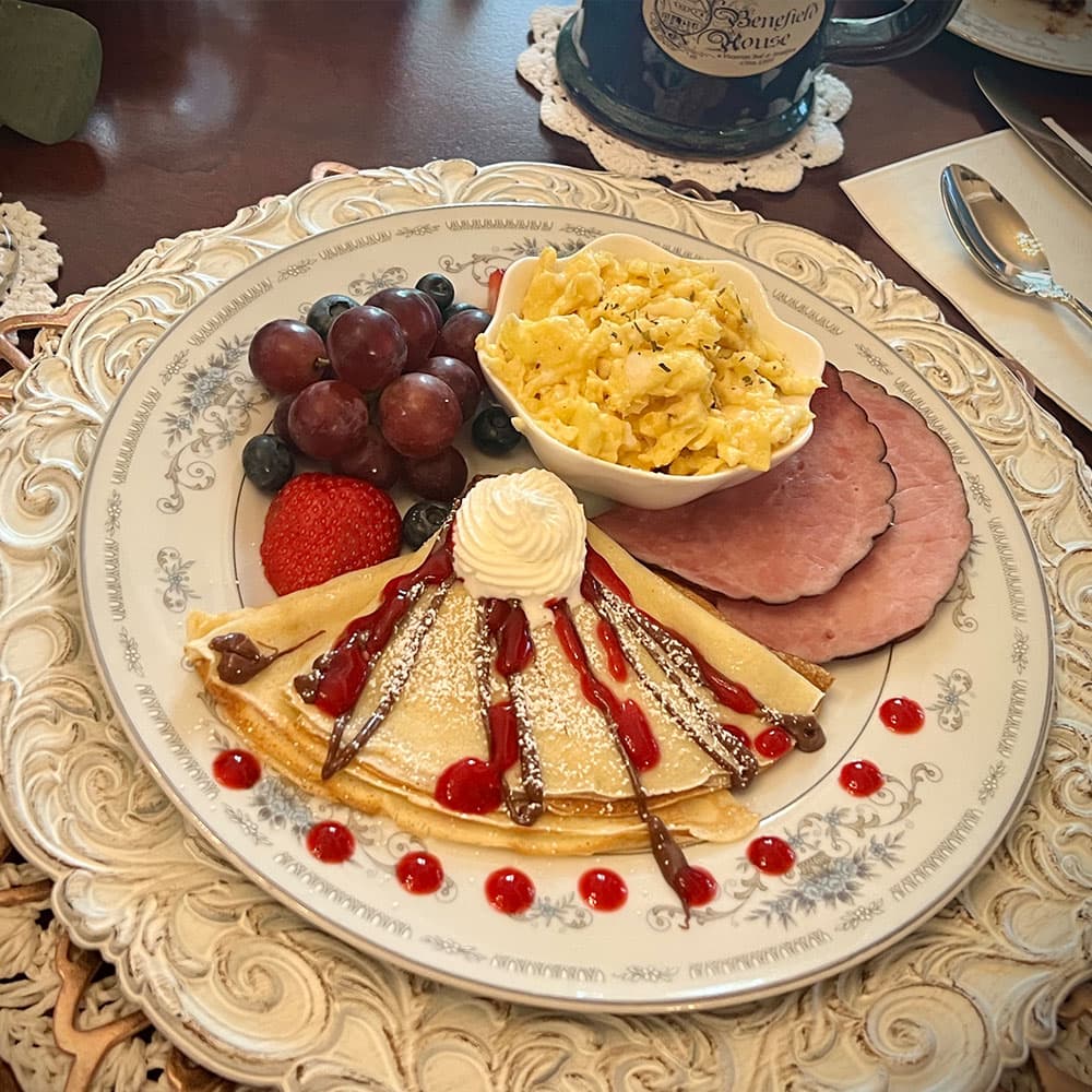 A beautifully plated breakfast featuring crepes with chocolate and berry sauce, scrambled eggs, ham, and assorted fruits.