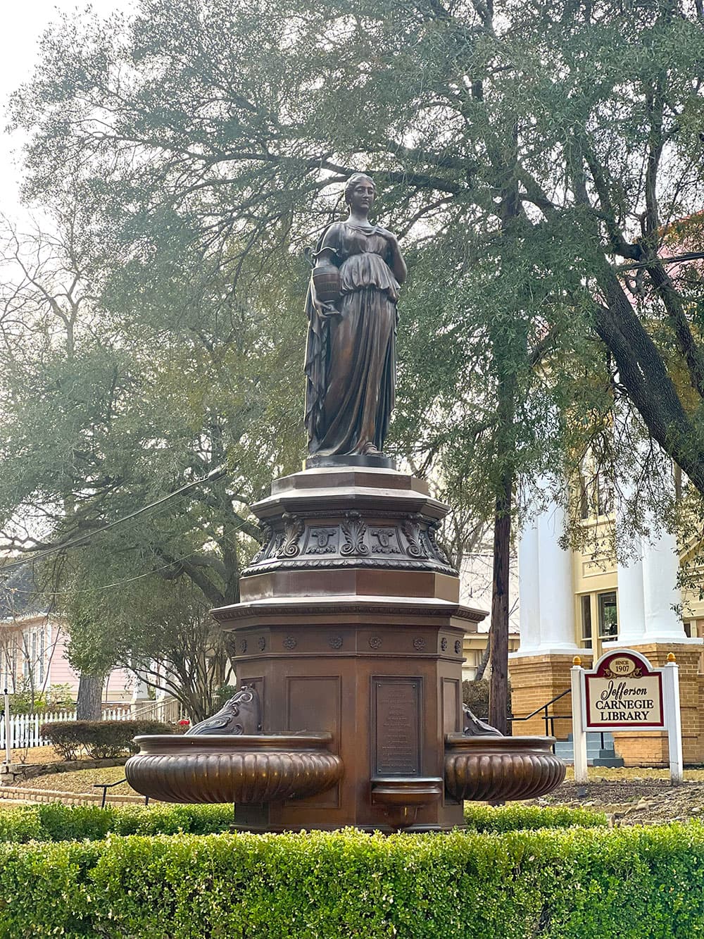 A bronze statue of a woman holding a book stands atop a decorative fountain, surrounded by greenery and trees.